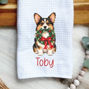 May include: A white waffle-textured kitchen towel featuring a printed illustration of a tricolor Corgi dog holding a green Christmas wreath with a red bow in its mouth, with the name Toby printed in red text below the dog.