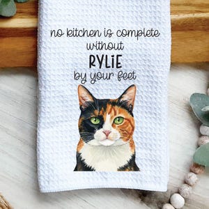 May include: White kitchen towel with a textured surface. The towel features a watercolor portrait of a calico cat with green eyes. Text reads "no kitchen is complete without RYLIE by your feet."