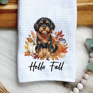 May include: A white waffle-textured kitchen towel featuring a graphic of a black and tan puppy wearing a plaid bandana, surrounded by orange pumpkins and autumn leaves, with the black text Hello Fall printed underneath the illustration.