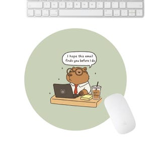 May include: A round, light green mousepad featuring a cartoon capybara wearing glasses and a tie, working on a laptop. The mousepad has the text "I hope this email finds you before I do". A white mouse and keyboard are also visible.