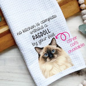 May include: White kitchen towel with a textured waffle weave. The towel features a Ragdoll cat illustration with blue eyes and the text "no kitchen is complete without a RAGDOLL by your feet." Additional text says "other cats available."