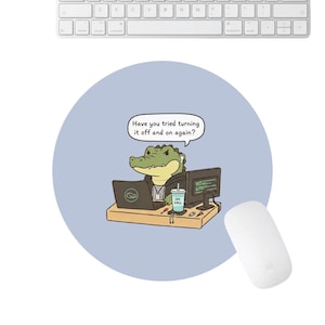 May include: A round, light blue mousepad featuring a cartoon alligator in a suit working on a laptop and desktop computer. The alligator has a speech bubble that says, "Have you tried turning it off and on again?" A white mouse and keyboard are also visible.