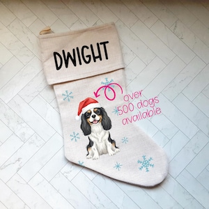 May include: A beige Christmas stocking with the name "DWIGHT" in black block letters. The stocking features a Cavalier King Charles Spaniel wearing a Santa hat, surrounded by blue snowflakes. Text reads "over 500 dogs available."