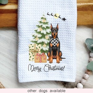 May include: A white waffle-textured kitchen towel featuring a Doberman wearing a black and white buffalo plaid bandana, a decorated Christmas tree, wrapped gifts, a silhouette of Santa's sleigh, and the text Merry Christmas.