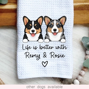 May include: A white waffle-textured kitchen towel featuring a graphic of two Corgi dogs. The text reads, Life is better with Remy & Rosie, followed by a small heart symbol. The dogs are illustrated in black, white, and tan colors.