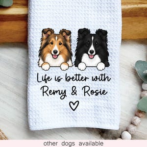 May include: A white waffle-textured kitchen towel featuring a graphic of two dogs, one sable and white and one black and white, with the text "Life is better with Remy & Rosie" and a small black heart symbol printed in the center of the fabric.