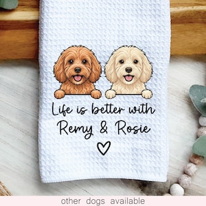 May include: A white waffle-textured kitchen towel featuring a graphic of two dogs, one brown and one cream, with the text "Life is better with Remy & Rosie" printed in black, accompanied by a small black heart symbol centered below the names.