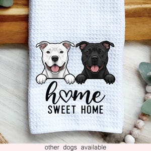 May include: White decorative kitchen towel featuring a smiling white and black dog illustration. The towel has the words "home sweet home" in black script. The towel is made of a textured material.