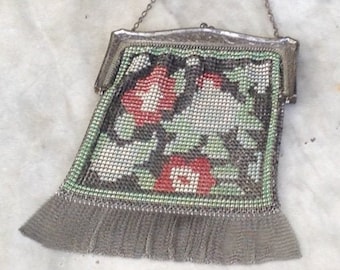 May include: Vintage silver mesh purse with a floral design in red, green, and white. The purse has a silver chain handle and a fringe at the bottom. The floral pattern is detailed and intricate, with a classic, antique aesthetic.