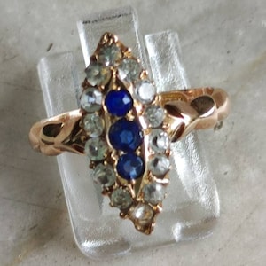 May include: A gold ring with a marquise-shaped setting featuring three blue sapphires surrounded by smaller clear stones.