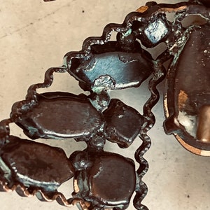 K&ouml;nnte beinhalten: Nahaufnahme einer Vintage-Brosche mit einem Cluster aus dunkelbraunen, facettierten Steinen, die in einem kupferfarbenen Metallrahmen gefasst sind. Die Steine sind in einer dreieckigen Form angeordnet und haben ein leicht abgenutztes Aussehen.
