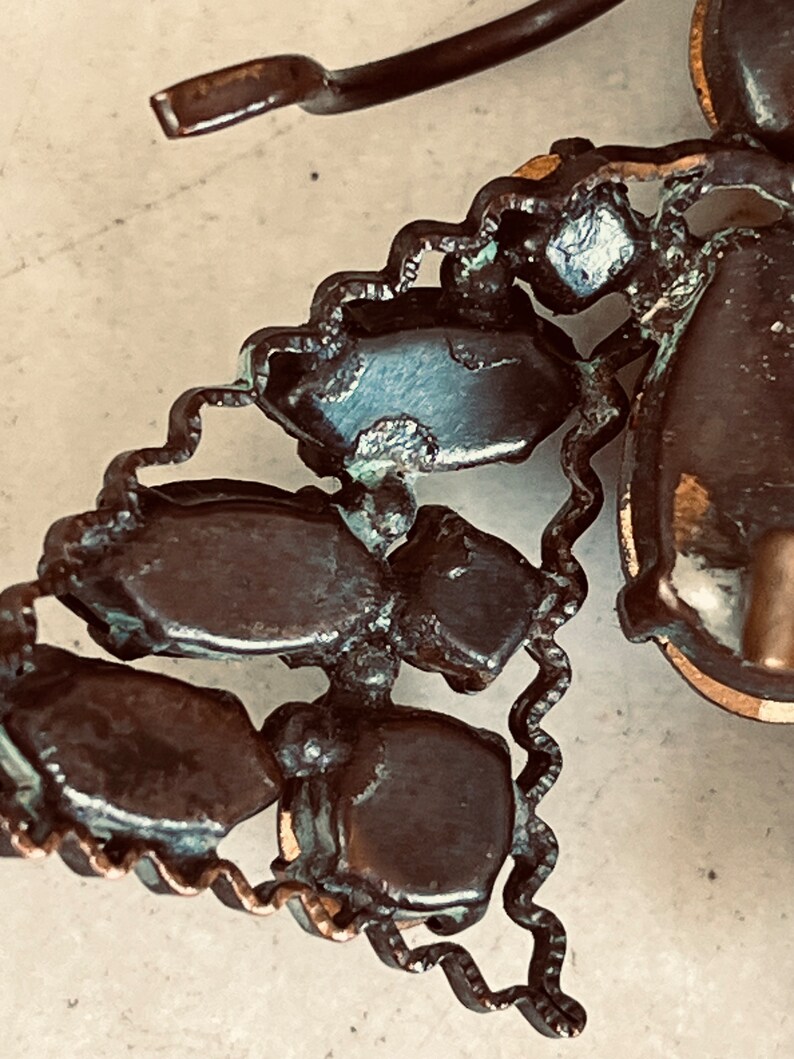 K&ouml;nnte beinhalten: Nahaufnahme einer Vintage-Brosche mit einem Cluster aus dunkelbraunen, facettierten Steinen, die in einem kupferfarbenen Metallrahmen gefasst sind. Die Steine sind in einer dreieckigen Form angeordnet und haben ein leicht abgenutztes Aussehen.