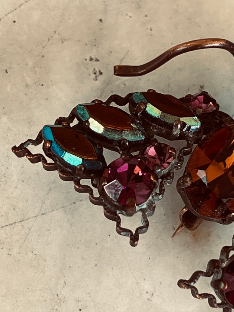 K&ouml;nnte beinhalten: Eine Vintage-Brosche mit einem dekorativen Design aus schillernden blauen und violetten Glassteinen und einer goldfarbenen Metallnadel.