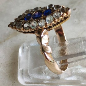 May include: A gold ring with a large, eye-shaped setting. The setting is filled with small, clear stones and three larger, blue stones. The ring band is plain and smooth.