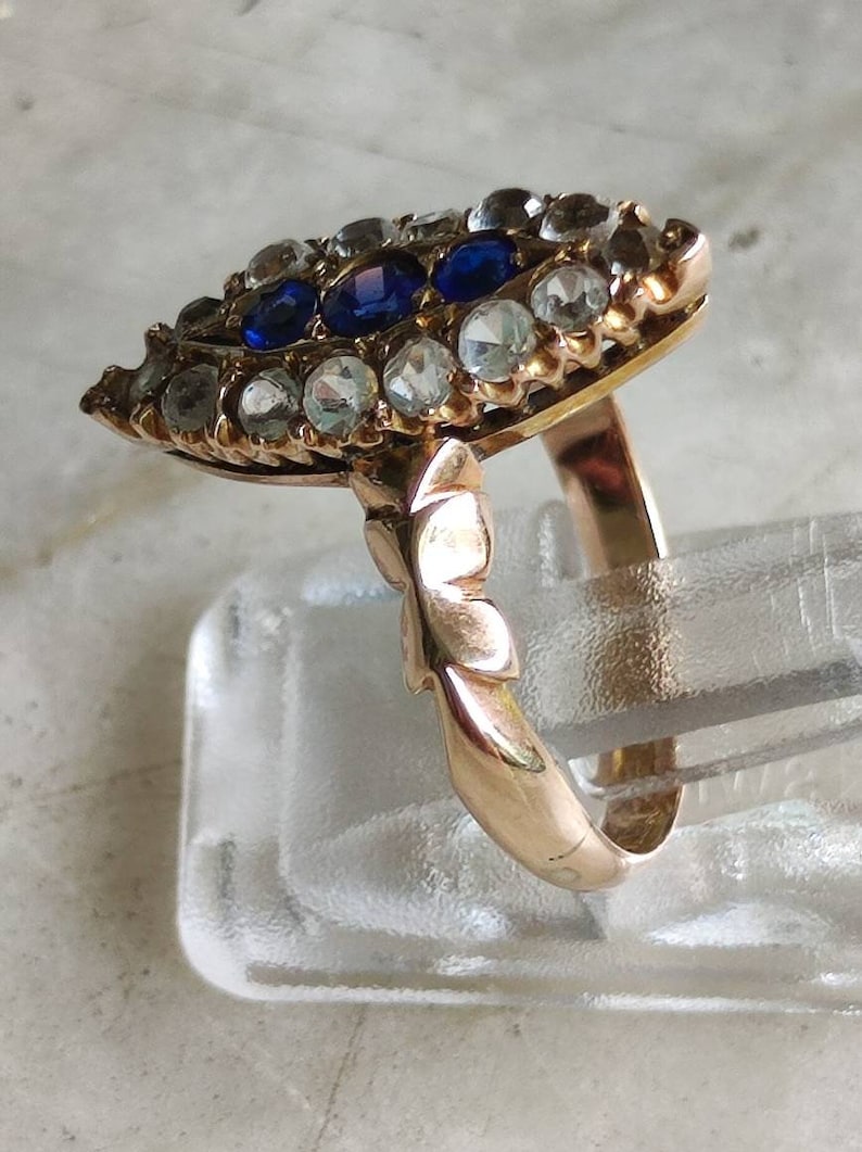 May include: A gold ring with a large, eye-shaped setting. The setting is filled with small, clear stones and three larger, blue stones. The ring band is plain and smooth.