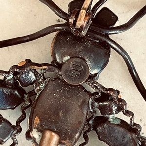 K&ouml;nnte beinhalten: Die R&uuml;ckseite einer Vintage-Brosche in Form einer stilisierten Spinne. Die Brosche ist aus Metall und hat eine schwarze Oberfl&auml;che. Die Spinne hat einen runden K&ouml;rper mit einem schwarzen Stein in der Mitte. Die Beine der Spinne sind aus Draht und mit schwarzen Steinen besetzt. Die Brosche hat eine Nadel auf der R&uuml;ckseite, um sie an Kleidung zu befestigen.
