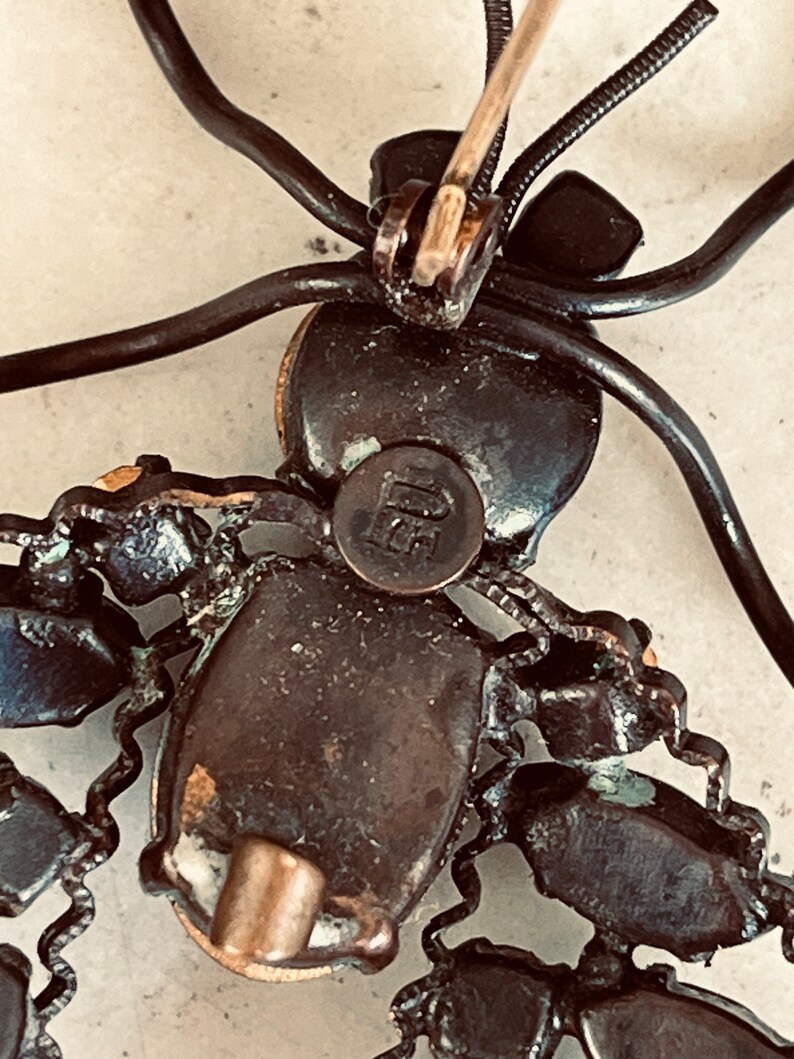 K&ouml;nnte beinhalten: Die R&uuml;ckseite einer Vintage-Brosche in Form einer stilisierten Spinne. Die Brosche ist aus Metall und hat eine schwarze Oberfl&auml;che. Die Spinne hat einen runden K&ouml;rper mit einem schwarzen Stein in der Mitte. Die Beine der Spinne sind aus Draht und mit schwarzen Steinen besetzt. Die Brosche hat eine Nadel auf der R&uuml;ckseite, um sie an Kleidung zu befestigen.