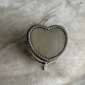 May include: A silver heart-shaped trinket box with a decorative border and a textured interior. The box has a hinged lid and a small foot on the bottom.
