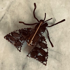 K&ouml;nnte beinhalten: Eine Vintage-Brosche in Form eines Schmetterlings, mit einem dunkelbraunen Metallrahmen und schwarzen Strasssteinen. Die Brosche hat eine Stecknadel- und Verschluss-Verschluss auf der R&uuml;ckseite.