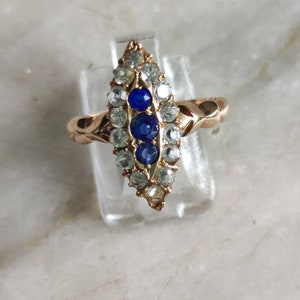May include: A gold ring with a marquise-shaped setting featuring a row of blue sapphires surrounded by smaller clear stones.