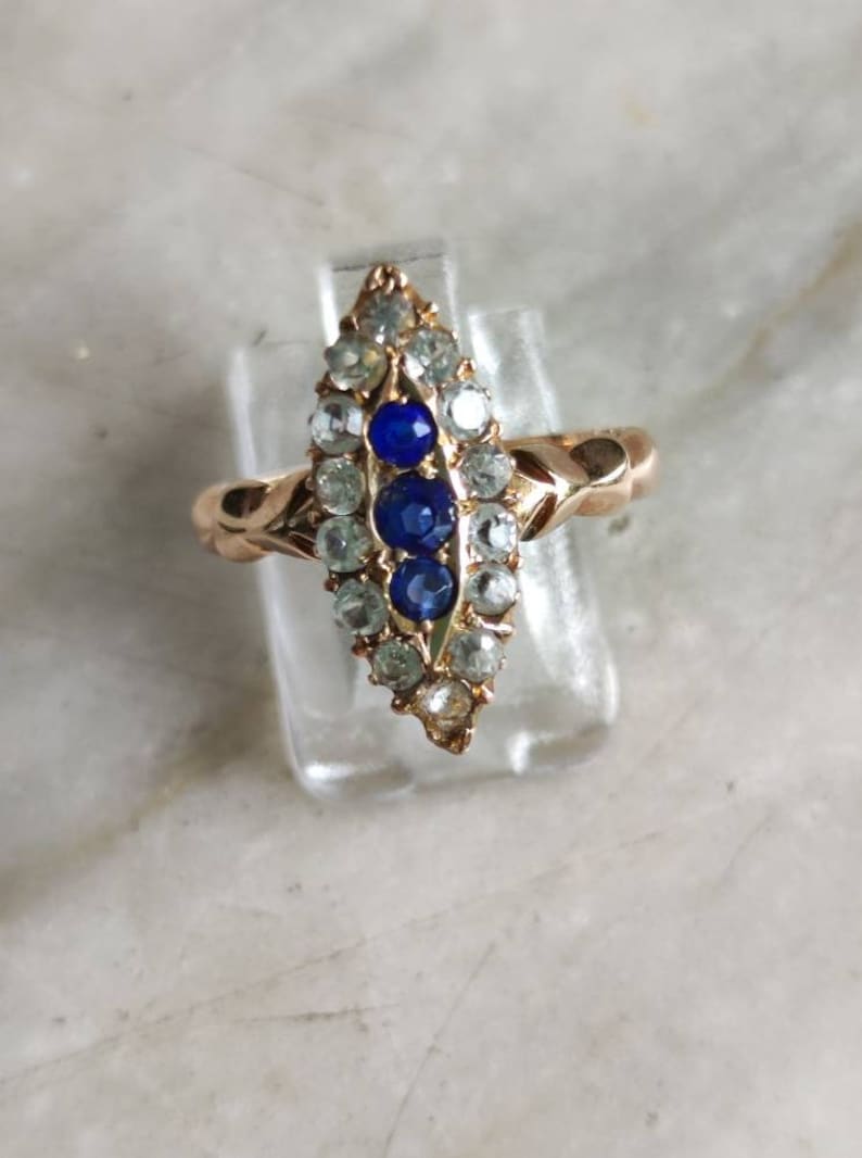 May include: A gold ring with a marquise-shaped setting featuring a row of blue sapphires surrounded by smaller clear stones.