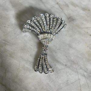 May include: A silver-coloured brooch in a fan design, embellished with clear rhinestones. The brooch features a central rectangular section and a tassel-like bottom, all set with sparkling stones. The design evokes vintage jewellery.