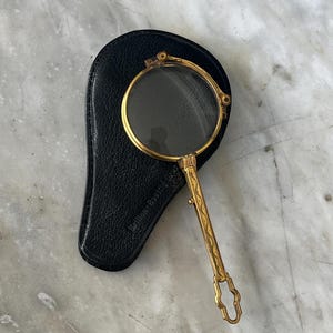 Op de afbeelding: Een vintage vergrootglas in goudkleur met een ronde lens, op een zwart leren etui. De handgreep heeft een sierlijk ontwerp. Op de etui staat "Bellen-Boeritz".