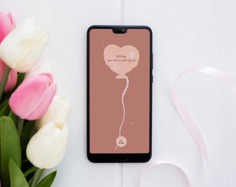 May include: A smartphone displays a heart-shaped balloon graphic with the text "Darling you are a work of art." The phone is surrounded by pink and white tulips and a pink ribbon on a white wooden surface. The phone's screen has a muted pink background.