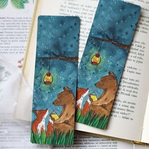 Può includere: Due segnalibri illustrati con un orso e una volpe che leggono un libro sotto un cielo stellato. Una lanterna è appesa a un ramo sopra di loro. I segnalibri sono stampati su cartoncino e hanno uno stile ad acquerello.