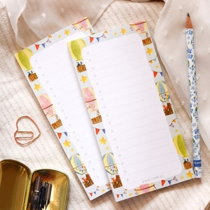 May include: Two white lined notepad pads with a colourful hot air balloon pattern. The pattern includes yellow, blue, red, and green hot air balloons with stars and flags.