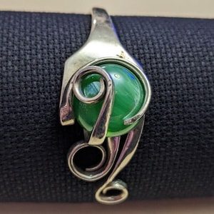 May include: A silver pendant with a bright green orb. The pendant has a swirling, decorative design that holds the green sphere. The metal has a polished finish, contrasting with the smooth, reflective orb. A unique piece of jewellery.