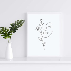 May include: A white framed print with a black line drawing of a face with a flower growing out of the eye. The print is on a white shelf with a white vase and green leaves.