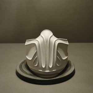 May include: A white, 3D printed mask with a geometric design. The mask is on a black circular base.