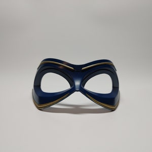 May include: A navy blue eye mask with gold trim. The mask has a curved shape with two eye openings and is likely made of plastic or a similar material. The gold trim accents the edges of the mask.