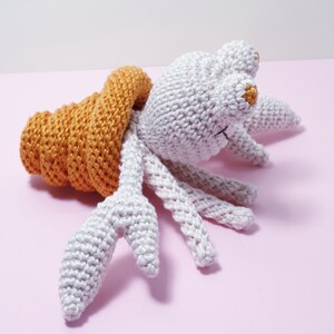 Peut inclure: Un jouet homard crocheté à la main en gris clair et orange. Le homard a un corps gris clair, plusieurs pattes et des pinces orange. Le jouet est posé sur une surface rose.