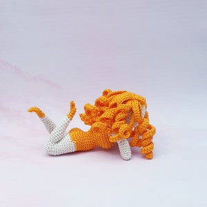 Peut inclure: Une poupée en crochet avec des cheveux orange et un corps blanc et orange allongée sur le côté.