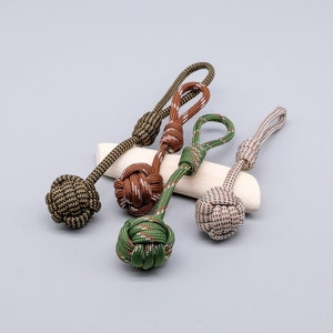 Pode incluir: Quatro chaveiros de corda coloridos com bolas de nó. Os chaveiros são feitos de corda verde, marrom e bege.