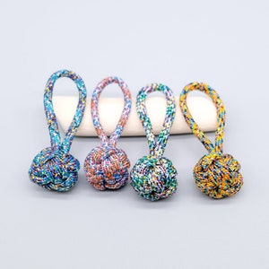 Può includere: Quattro portachiavi con nodi colorati, annodati a mano. Ogni portachiavi presenta un nodo rotondo con un anello, realizzato con un mix di cordoncini blu, viola, arancioni e verdi. Poggiano su una superficie bianca e curva su uno sfondo grigio chiaro.