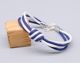 Op de afbeelding: Een nautische armband met een knoopontwerp. De armband is gemaakt van witte en marineblauwe koorden, met een zilverkleurige sluiting. De armband wordt gepresenteerd op een klein houten blok tegen een lichtgrijze achtergrond.
