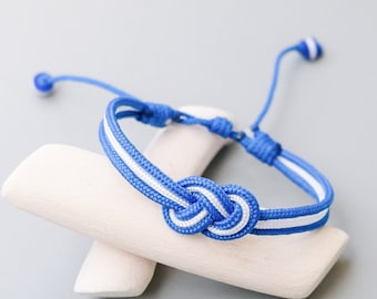 Op de afbeelding: Een blauw en wit gestreept vriendschapsarmband met een knoop in het midden. Het armband wordt gepresenteerd op een wit houten oppervlak.