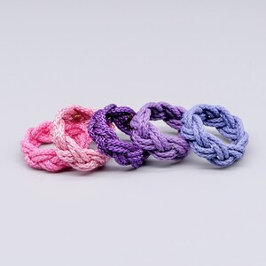 Könnte beinhalten: Vier geflochtene Ringe in Rosa- und Lilatönen. Die Ringe sind aus einem weichen, flexiblen Material gefertigt und perfekt, um jedem Outfit einen Hauch von Farbe zu verleihen.