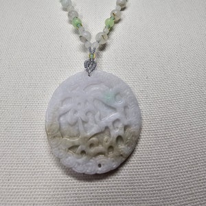 May include: A white jade necklace with a round pendant featuring intricate carvings. The pendant is a light green color and has a hole in the center. The necklace is made of white and green beads.