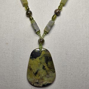 May include: A necklace featuring a large, irregular, olive-green and black stone pendant suspended from a strand of alternating olive-green rectangular beads, small dark brown spherical beads, and tiny translucent green accent beads.