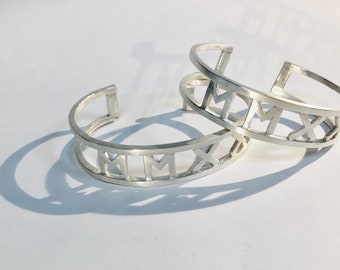 May include: Two silver bracelets with a Roman numeral design. The bracelets are open and have a simple, minimalist style.