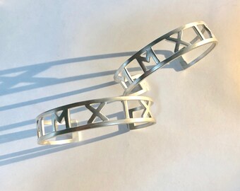 May include: Two silver bracelets with Roman numeral cutouts. The bracelets are stacked on top of each other.