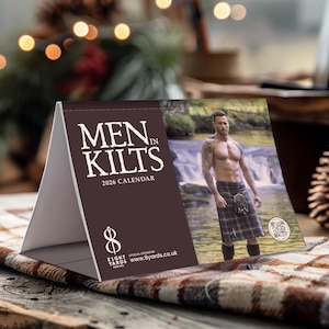 Peut inclure: Calendrier 2026 intitulé "MEN in Kilts" avec un homme en kilt près d'une cascade. Le calendrier a un fond marron avec du texte blanc et un logo. Le calendrier est présenté sur une surface en bois.