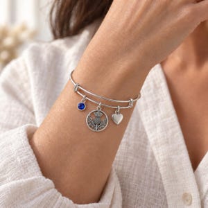 May include: A silver-toned expandable wire bangle bracelet featuring three dangling charms: a circular pendant with a detailed thistle design, a small smooth heart, and a single round blue crystal stone set in a silver-toned metal frame.