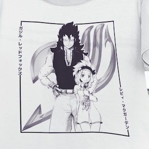 May include: White t-shirt with a black and white illustration of two anime characters. The characters are standing next to each other and are surrounded by a black and white symbol. The text "ガジル・レッドフォックス" and "レヴィ・マグガーデン" is written in Japanese on the shirt.