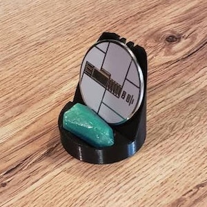 May include: A black 3D printed stand with a round base and a circular top. The stand holds a round, silver-colored object with a black and white design. A teal-colored object is resting in the base of the stand.