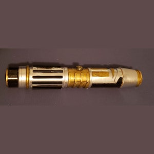 Peut inclure: Un sabre laser argenté et doré avec des accents noirs. Le sabre laser a une surface texturée et une poignée noire.
